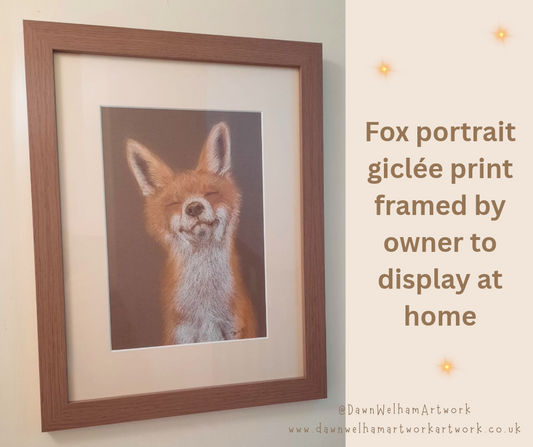 Framed fox portrait print with a warm brown wood frame and pale cream mount, shown hanging on a light wall. The artwork depicts a fox face on a muted brown background.